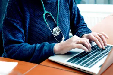 Foto van handen die druk bezig zijn op laptop, om de hals van de persoon hangt een stethoscoop