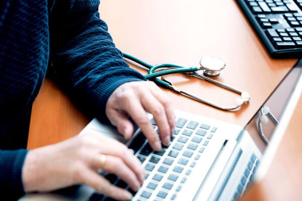 Foto van handen die op een laptop bezig zijn, naast de laptop ligt een stethoscoop