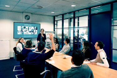 Foto van collega's bij elkaar aan tafel, luiterend naar een presentatie